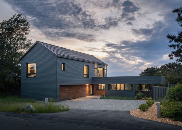 Exterior view of a contemporary two-storey home at dusk, featuring dark horizontal cladding, a pitched metal roof, warm-lit windows, an integrated lower-level garage, and a single-storey connecting wing. The house sits within a landscaped suburban setting with a gravel driveway and planting.
