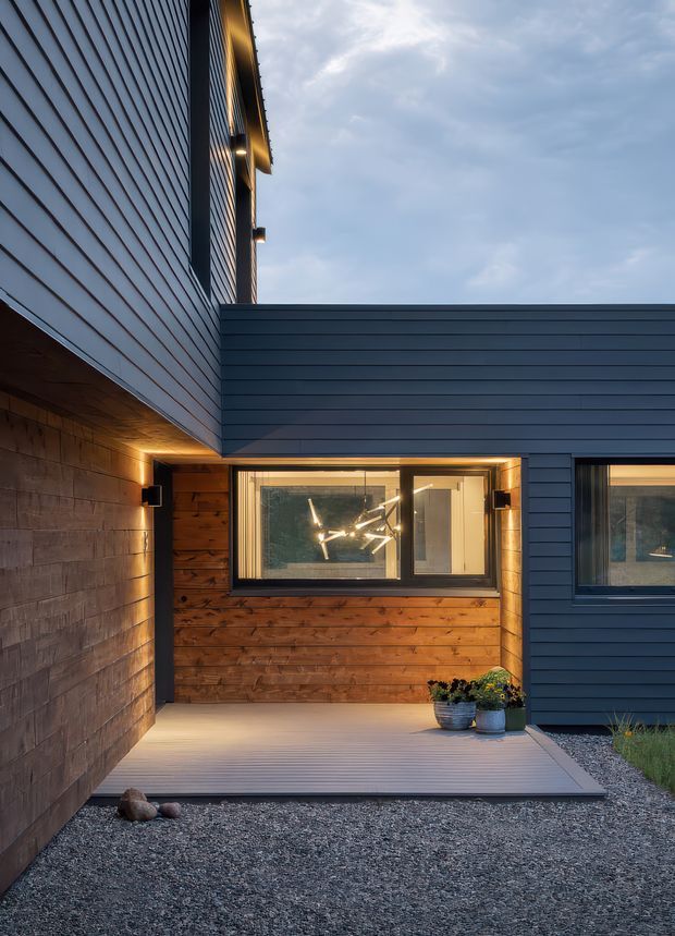 Exterior architectural photograph of a contemporary home entry at dusk. The composition shows dark horizontal cladding, a projecting flat-roofed volume, warm timber wall lining beneath a sheltered porch, integrated exterior lighting, windows, and a pale composite or timber-look deck leading through a gravel forecourt. A partially visible interior pendant light is seen through the glazing.