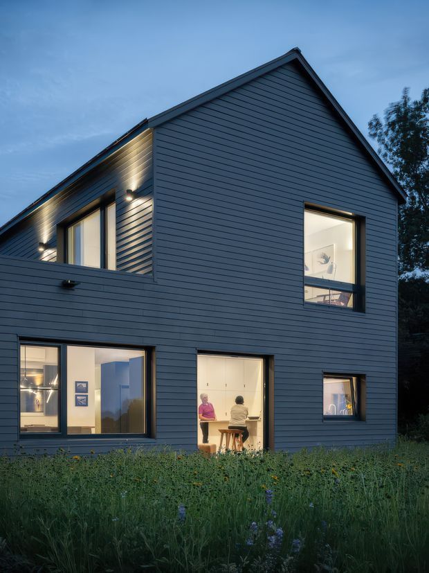 Exterior dusk view of a contemporary two-storey home with a steep gabled roof and dark horizontal weatherboard cladding. Large windows and glazed openings reveal warmly lit interior spaces, including a kitchen or dining area, while exterior wall lights create a welcoming evening atmosphere. The house is set against a planted garden landscape.