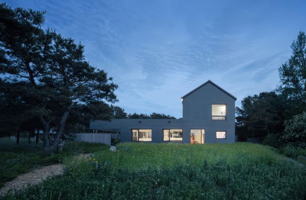A contemporary two-part residential house photographed at dusk, set within a leafy landscaped site. The main two-storey volume has a steep simple gable roof and dark horizontal cladding, with warm light glowing from the windows and ground-floor openings. A lower single-storey wing extends to the left, while a gravel path crosses a meadow-like garden in the foreground and mature trees frame the building.