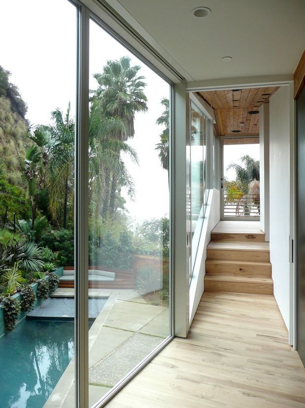 A bright contemporary interior corridor and stairway lined with full-height glazing overlooking a lush tropical garden and narrow pool. The hallway has pale timber flooring, matching timber stair treads, white walls, slim aluminium-framed glass doors and windows, and a warm timber-lined ceiling over the stair landing. A balcony railing and garden are visible beyond the glazed opening.