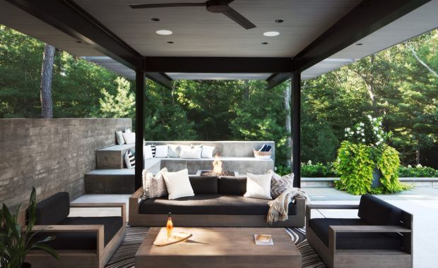 A sophisticated covered outdoor lounge with a dark timber or metal structural frame, light slatted ceiling, and a ceiling fan. The space includes built-in concrete seating along the rear and side walls, a central dark upholstered sofa, matching armchairs, a low timber coffee table, patterned rug, cushions, and a small fire feature. It opens directly onto a lush wooded garden, creating a refined indoor-outdoor entertaining area.
