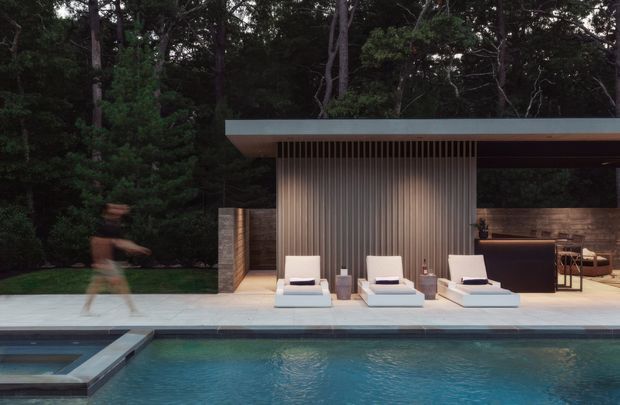 A refined modern poolside entertaining pavilion set within a wooded landscape. The single-storey structure has a broad flat roof with a deep overhang, dark slender framing, a vertical ribbed feature wall, and warm horizontal timber-clad walls. Three white loungers sit on a pale paved terrace beside a swimming pool, with a partially enclosed outdoor bar and seating area visible at right. A blurred person passes along the left side, conveying movement in an otherwise calm, moody evening setting.