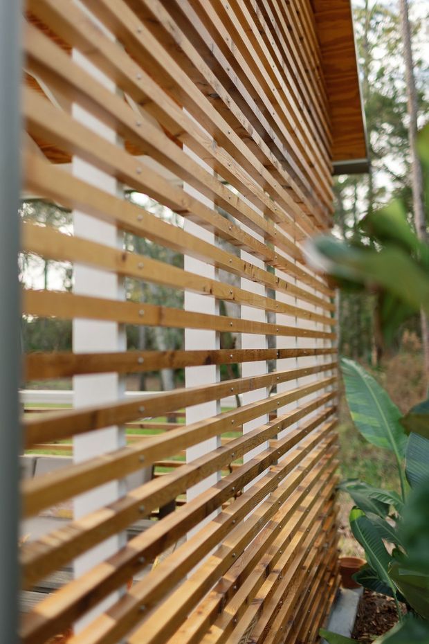 Close-up exterior architectural view of a long horizontal timber slat screen or louvred facade. Warm natural wood battens are fixed to white structural posts, creating a layered semi-transparent privacy and sun-shading element beside a planted outdoor setting. A timber roof overhang is visible at the top, with greenery seen through and beside the screen.
