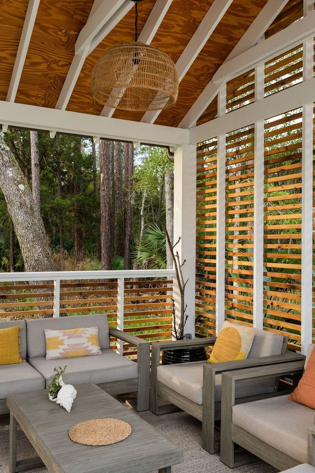A covered outdoor lounge room or screened patio set within a wooded landscape. The space has a pitched ceiling with exposed white-painted rafters and warm plywood panels, horizontal timber privacy screens, white structural framing and railings, and grey wood-framed upholstered seating arranged around a low coffee table. A woven rattan pendant light and natural-textured accessories add warmth to the relaxed outdoor setting.