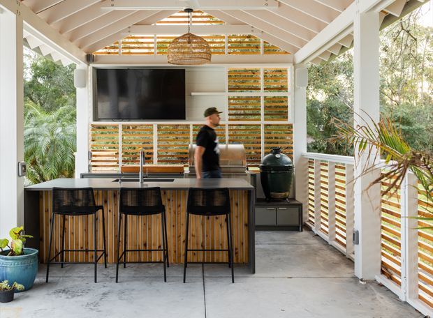A bright, open-sided covered outdoor entertaining area with a pitched timber roof, integrated outdoor kitchen and barbecue, and a bar island with three stools. Horizontal timber privacy screens line the rear and side walls, while white timber posts and railings frame views of lush vegetation. A woven pendant light, wall-mounted television, concrete floor, and built-in cooking appliances complete the relaxed outdoor living space.