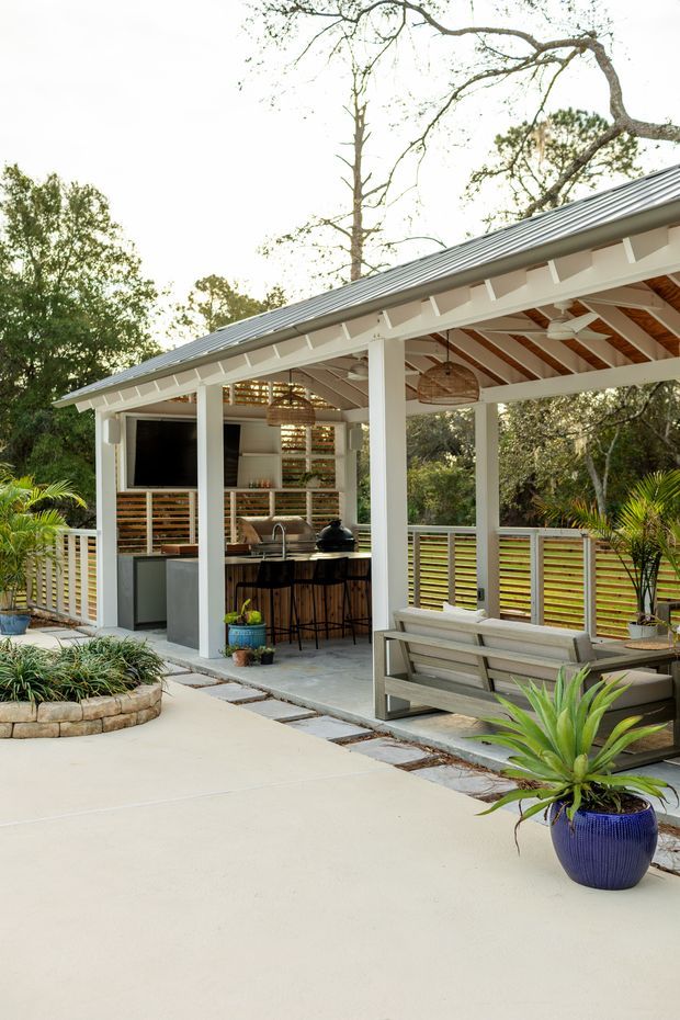 A landscaped outdoor entertaining pavilion with a covered open-air kitchen and bar, timber-framed gable roof, white structural posts and beams, horizontal timber privacy screens, ceiling fans, woven pendant lights, and a built-in bench. The space opens onto a light concrete patio surrounded by mature greenery and potted tropical plants.