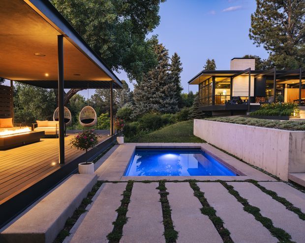 Twilight exterior view of a contemporary, multi-level residence set in a landscaped garden. A covered outdoor living pavilion with timber decking and integrated seating occupies the left side, while the main house features dark-framed glazing, light horizontal cladding and a low-pitched roof. A small illuminated swimming pool, concrete retaining walls and large concrete pavers with planted joints form the foreground.