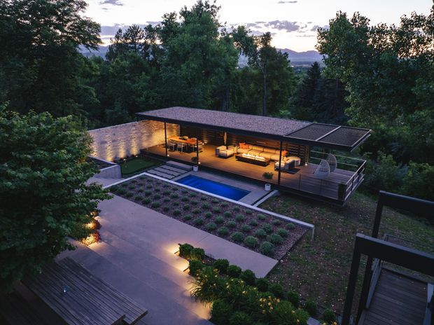 Aerial dusk view of a low-profile modern pavilion-style house set within a wooded landscape. The building has broad flat or very low-pitched roof planes, extensive open glazing, warm illuminated timber-lined interiors, and a large outdoor deck. A rectangular swimming pool, paved terrace, landscaped planting beds, and timber pathways surround the residence, with wooded hills visible beyond.