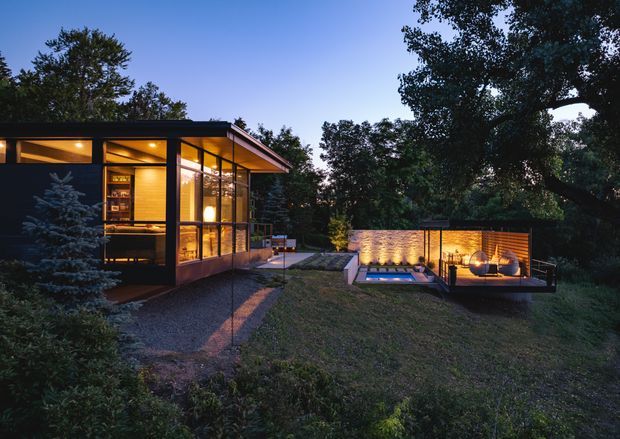 Twilight exterior view of a low, modern pavilion-style residence set among mature trees. The glazed main volume has dark slender framing, warm interior lighting, and a broad shallow roof. A gravel path leads toward a small illuminated pool or water feature and a separate timber-clad outdoor pavilion with built-in seating and a circular fire feature.