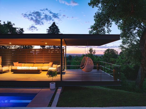 A modern, open-sided outdoor entertaining pavilion at dusk, set among mature trees. The covered timber deck contains built-in lounge seating arranged around a linear outdoor fire feature, horizontal timber screening, recessed ceiling lights, and a suspended egg chair. A glass or metal balustrade borders the deck, with a swimming pool and landscaped lawn in the foreground.