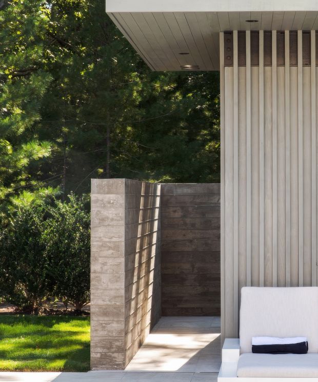 A close-up exterior architectural view of a contemporary pavilion or house corner set within a leafy garden. The building features a deep flat roof overhang with a pale panelled soffit, vertical light-coloured cladding, and a recessed outdoor passage enclosed by textured concrete walls. A built-in or outdoor lounge chair is partially visible at the right edge. Sunlight creates strong shadows across the pale paving and timber-toned wall surfaces, with mature greenery surrounding the structure.