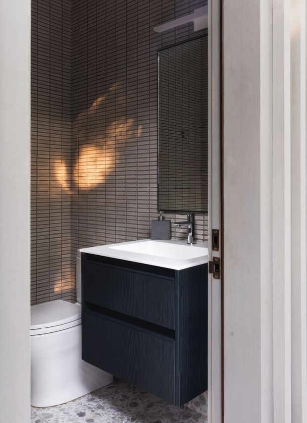 A compact contemporary bathroom viewed through an open door, featuring dark grey linear mosaic wall tiles, a black fluted floating vanity with a white integrated basin, chrome wall-mounted tapware, a tall rectangular mirror, and a white toilet. The floor is finished in small irregular grey stone-like tiles, creating a refined, high-contrast scheme.
