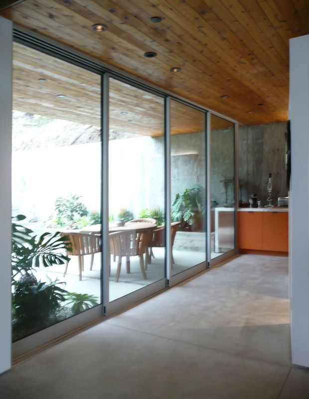 Interior view of a modern dining and kitchen area connected to a planted courtyard through extensive floor-to-ceiling glazing. The space features a warm timber-lined ceiling with recessed downlights, polished concrete flooring, slim dark metal-framed glass panels, exposed concrete walls, and bright orange-red kitchen cabinetry.