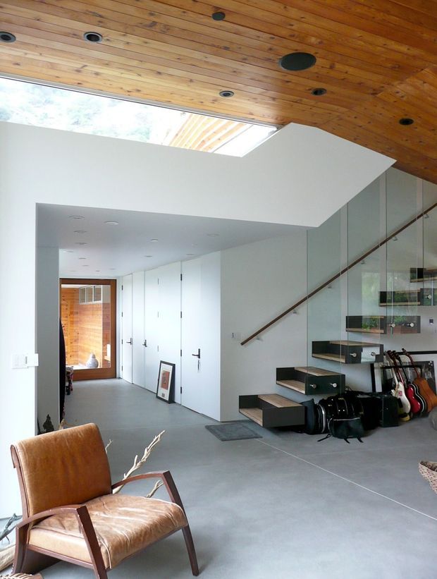 A modern open-plan interior featuring a floating staircase with dark treads and integrated drawer-like storage, a glass balustrade, and guitars displayed beneath the stairs. The space has a polished concrete floor, crisp white walls and doors, and a warm timber-lined ceiling with recessed spotlights. A large skylight or clerestory opening brings in daylight, while a leather lounge chair occupies the foreground.