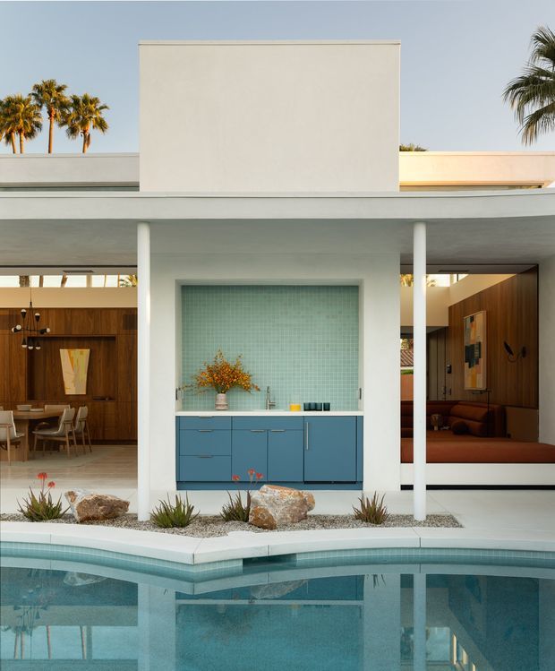 A symmetrical mid-century-inspired residential courtyard centered on a turquoise swimming pool. A white flat-roofed pavilion with slender columns frames an outdoor wet bar or kitchenette with blue cabinetry and a pale aqua mosaic-tile backsplash. Openings on either side reveal warm wood-paneled dining and lounge areas. Desert-style planting, gravel, rocks, and palms reinforce the warm modernist setting.