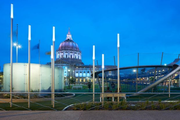An exterior urban architectural scene at dusk, featuring a monumental domed civic building illuminated in the background. The foreground contains a landscaped public space with tall slender light poles, curved paved paths, low planting, metal railings, and a large curved mesh or cable structure. The composition has a vivid blue evening sky and a contemporary civic landscape setting.