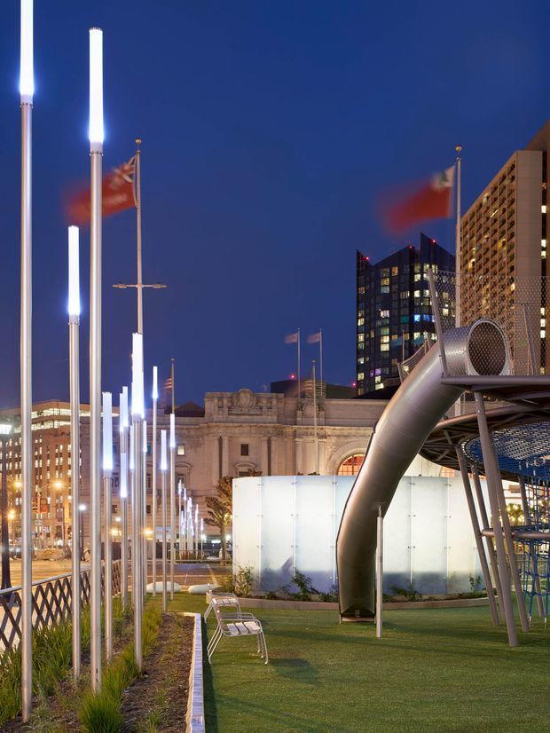 A dusk view of a landscaped urban public plaza with rows of illuminated flagpoles and linear light poles, lawn, benches, and a large sculptural tubular play or architectural structure. Historic stone architecture sits behind the plaza, alongside contemporary illuminated high-rise buildings and a translucent white pavilion.