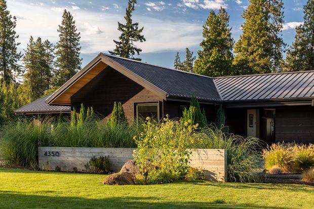 Exterior view of a low, single-storey timber-clad house set within a landscaped garden and surrounded by mature trees. The home has intersecting pitched gable roofs with dark standing-seam metal roofing, deep eaves, a recessed entry, and a broad timber-clad facade. Ornamental grasses, flowering plants, a concrete retaining planter, lawn, and boulders form the foreground landscape.