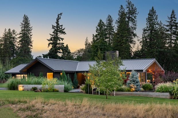 Single-storey contemporary house set within a landscaped, semi-rural wooded setting. The long low facade features dark horizontal timber cladding, a pale grey metal roof with multiple intersecting pitched sections, warm illuminated windows and entry doors, and extensive ornamental planting with grasses, shrubs, and young trees. The image is photographed from the front across a lawn at dusk, with tall conifers behind the property.