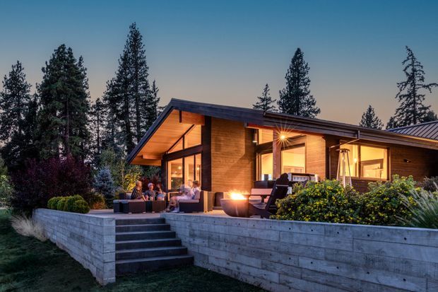 A single-storey contemporary timber-clad house nestled among tall trees, photographed at dusk. The home features a broad low-pitched roof with deep overhangs, extensive floor-to-ceiling glazing, warm interior lighting, and a raised concrete terrace with broad steps. Outdoor seating and a fire feature occupy the deck, surrounded by landscaped planting.