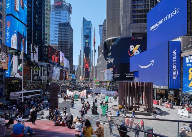 Wide urban streetscape viewed from elevated steps, showing a dense commercial city square lined with tall buildings, large digital billboards, retail and entertainment signage, pedestrians, and extensive paved public space. The scene is dominated by Times Square-style urban architecture and advertising displays in bright daylight.