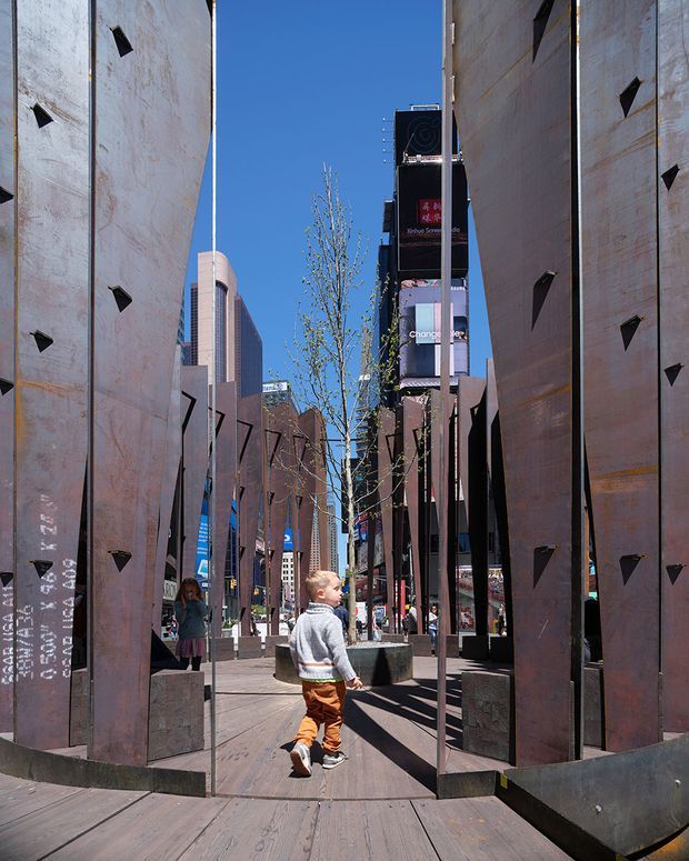 A sunny urban public plaza framed by tall rusted steel sculptural panels, with timber decking, concrete planters, young trees, pedestrians, and surrounding multi-storey commercial buildings. The strong vertical screens create a narrow passage through the city space, with bright blue sky and signage visible beyond.