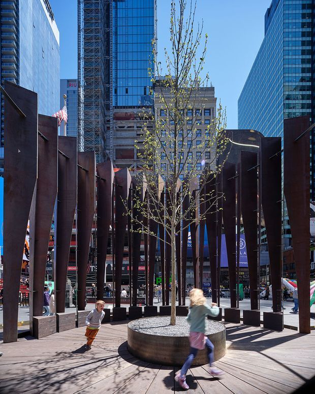 An urban public plaza framed by tall dark rusted-steel fins, with a young tree planted in a circular raised metal-edged planter at the centre. Timber decking forms the ground plane, while pedestrians move through the space. Dense glass high-rise buildings, construction scaffolding, signage and surrounding commercial facades create a dramatic downtown setting under clear daylight.