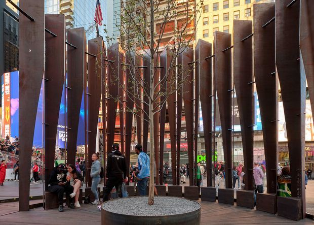 Urban public plaza framed by tall, rust-coloured steel fins arranged around a paved pedestrian space. Young trees grow from large circular planters filled with gravel, with integrated bench seating at the base of the screens. Surrounding high-rise buildings, retail signage, flags and pedestrians are visible beyond the installation. The image is an exterior civic or commercial public-space scene with a strong contemporary architectural intervention.