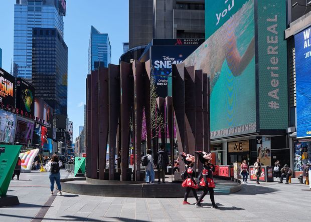 A bright daytime streetscape in a dense urban entertainment district, dominated by a sculptural dark-maroon architectural facade and entrance pavilion with tall angled fins. Large digital billboards and theatrical advertising surround the pedestrian plaza, with high-rise commercial buildings forming the backdrop and pedestrians crossing the paved public space.