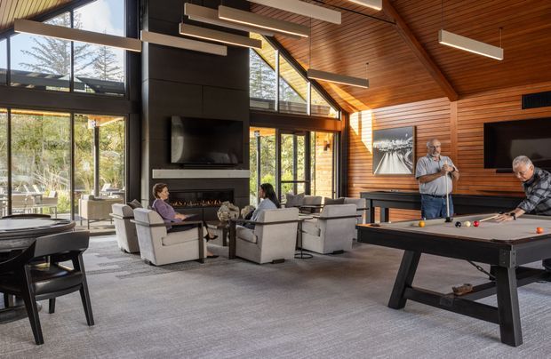 A spacious, double-height living and recreation room with a vaulted timber-lined ceiling, exposed structural beams, extensive floor-to-ceiling glazing, a central dark fireplace and media wall, lounge seating, a pool table, and a partial billiards table. The room has a warm timber interior, grey upholstered furniture, and contemporary linear pendant lighting.