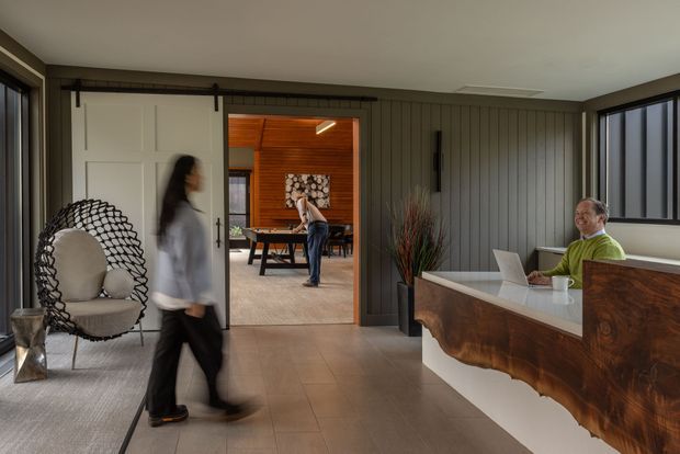 A sophisticated reception or entry space with a sculptural timber reception desk, a seated staff member, and a large sliding doorway leading into a warm timber-lined dining or meeting room. The room features dark vertical wall panelling, dark-framed glazing, a neutral tiled floor, and a statement woven lounge chair. The overall composition is contemporary with warm natural timber and refined hospitality styling.