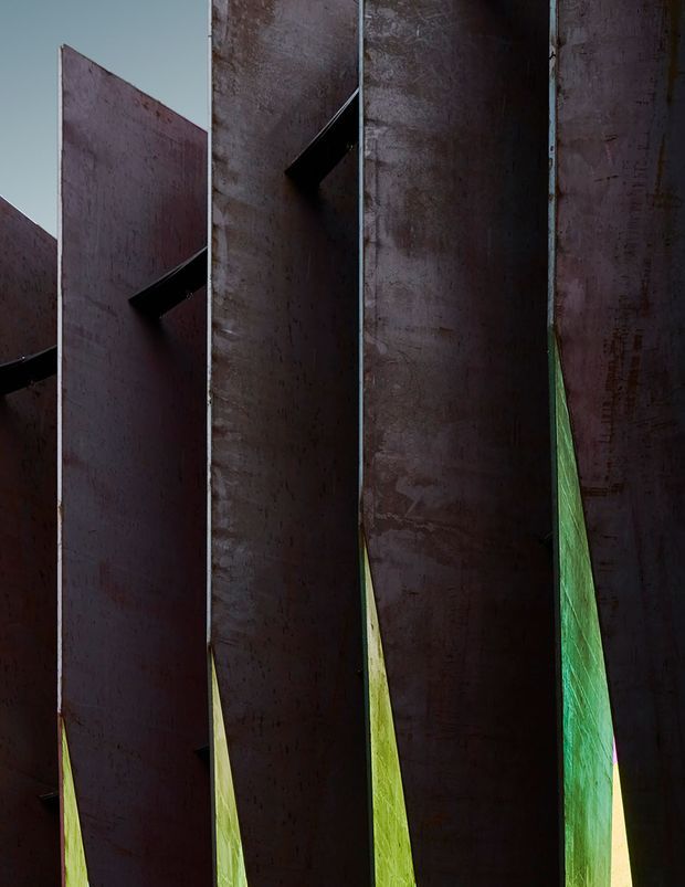 Close-up architectural view of a series of tall, narrow, weathered dark metal or concrete panels arranged in parallel, with sharp angular openings and vivid green foliage visible through the gaps. The composition is abstract, vertical, and highly textural, emphasizing strong geometric planes and deep shadows.