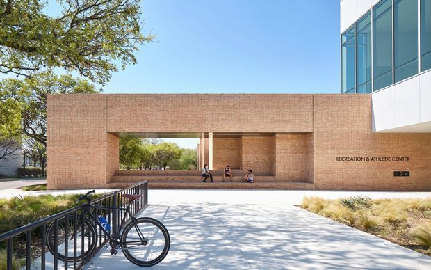 Exterior architectural view of a recreation and athletic center, featuring a long warm red-brick facade with a large recessed, framed opening and integrated bench seating. A contemporary white-and-glass building volume projects above at the right. The paved forecourt includes black metal bicycle railings and a bicycle, with established trees and ornamental landscaping around the building.