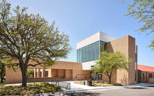 Exterior view of a contemporary multi-storey institutional or commercial building, featuring extensive floor-to-ceiling glazing, warm red-brown brick walls, pale metal-clad upper volumes, and a covered recessed entry. Mature trees, landscaped planting, paved walkways, and a black metal handrail frame the building.
