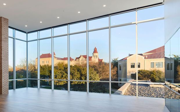 A bright, largely empty contemporary interior with a pale timber floor, exposed brick side wall, recessed ceiling lights, and extensive floor-to-ceiling glazing in slim dark frames. The glazing overlooks mature trees and nearby buildings with prominent red tiled roofs and a church-like tower. A reflective glass panel is visible at the right edge.