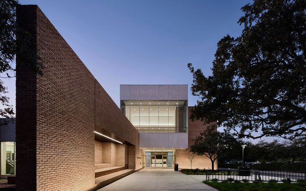 A contemporary institutional building exterior photographed at dusk, with long warm red-brown brick walls framing a glazed central entry pavilion. The entrance has expansive floor-to-ceiling glazing, a projecting upper box clad in pale panels, recessed linear lighting, and a broad paved approach. Mature trees and dark metal railings surround the forecourt.
