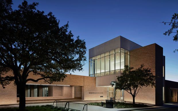 Dusk exterior view of a substantial contemporary brick building, likely a civic or institutional facility. The composition features a projecting two-storey glazed curtain-wall volume, flat roof forms, warm red-brown brick walls, landscaped entry planting, and a ramp with metal handrails. Mature trees frame the facade.