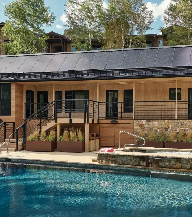 Exterior view of a contemporary timber-clad building beside a swimming pool. The structure has a long dark metal pitched roof with snow-retention detailing, black-framed glazing and railings, warm wood wall cladding, and a stone-clad raised pool edge with integrated water feature. Landscaped planters and a raised access deck are visible in front.