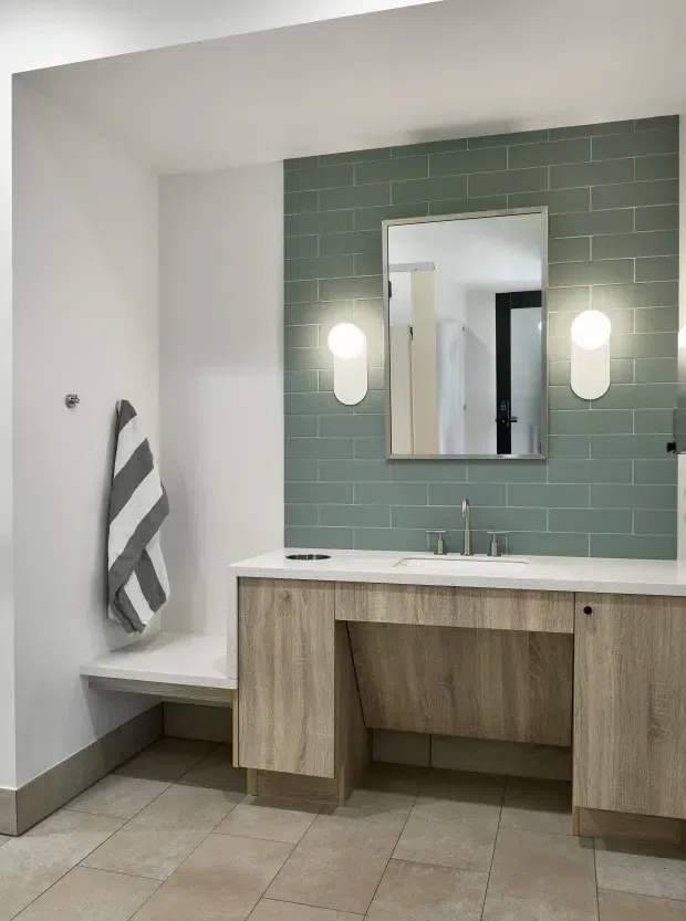 Contemporary bathroom vanity with a double-sink white benchtop, natural wood-look cabinetry, and a green-grey horizontally tiled feature wall. A large rectangular mirror and two rounded wall sconces sit above the vanity, while a built-in tiled bench and striped towel occupy the left side. Neutral floor tiles and white walls create a clean, understated interior.