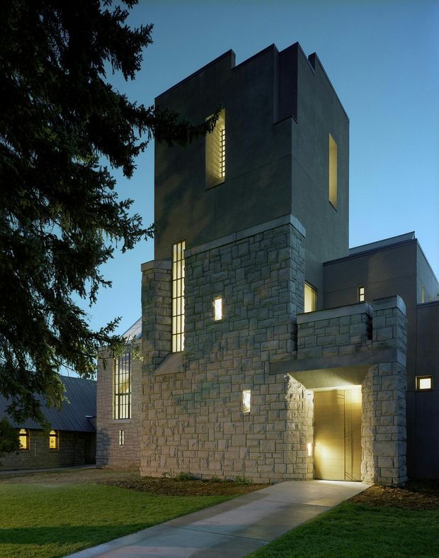 Dusk exterior view of a tall, sculptural contemporary building with dark rendered upper walls, extensive coursed grey stone masonry at the lower levels, narrow vertical illuminated windows, and a covered entrance reached by a concrete path. A steep dark metal roof and adjoining lower structures are visible, with mature evergreen trees and lawn framing the building.