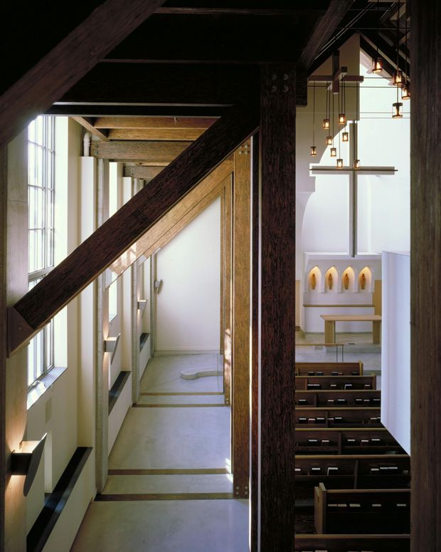 Interior view of a contemporary chapel or church, showing a narrow side aisle with tall vertical glazing, pale smooth walls and concrete flooring, contrasted with substantial dark exposed timber beams and posts. Through the structural opening, rows of dark timber pews face a simple altar area with a cross-like wall feature and suspended pendant lights. The composition emphasizes the dramatic exposed timber structure, strong vertical light, and sacred architectural space.