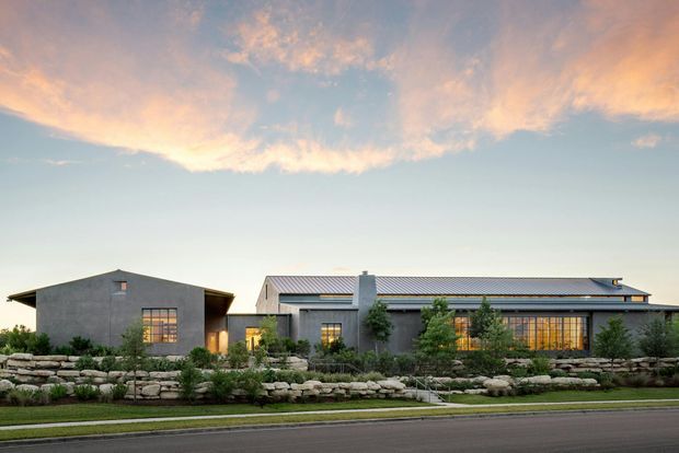 A wide exterior view of a low-profile contemporary residential building at dusk. The home is composed of simple rectangular volumes with dark grey rendered walls, metal pitched roofs, large gridded windows, and a central chimney-like vertical element. Layered natural stone retaining walls, structured planting, a paved path, and a street frontage occupy the foreground beneath a pastel sunset sky.