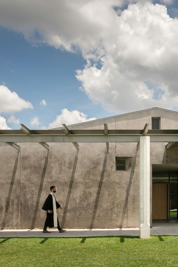 Exterior architectural photograph of a low-rise concrete or stucco building with a strong horizontal pergola or canopy, repeated angled supports, a high parapet roofline, and a recessed glazed entry. The weathered grey-beige wall surface and bright lawn are sharply lit beneath a dramatic blue sky with large clouds. A walking person provides scale.