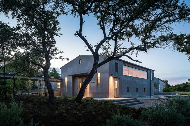 A contemporary single-storey concrete house set among mature trees and landscaped planting. The angular flat-roofed form has restrained openings, a large illuminated upper window, recessed entry, exterior steps, and dark metal-framed glazing. The image is taken from the landscaped exterior at dusk.