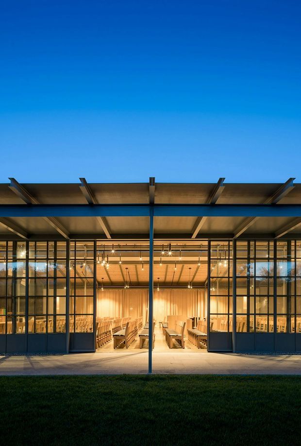 Exterior view of a contemporary glazed pavilion or hall at dusk, with a broad projecting flat canopy supported by dark slender steel columns and beams. Full-height gridded glazing and dark framed doors reveal a warmly illuminated interior arranged with rows of wooden seating, suggesting an auditorium, chapel, lecture hall, or community gathering space. The facade is set behind a paved forecourt and lawn, with a strong contrast between the deep blue sky and amber interior lighting.