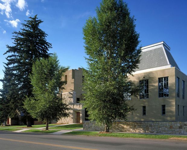 Street-level exterior view of a substantial multi-storey building partly screened by mature trees. The facade combines pale masonry walls with extensive stonework at the base and a prominent steep mansard-style roof section with grey shingle-like roofing. Tall divided-pane windows and a low stone boundary wall face the landscaped verge and roadway under a clear blue sky.