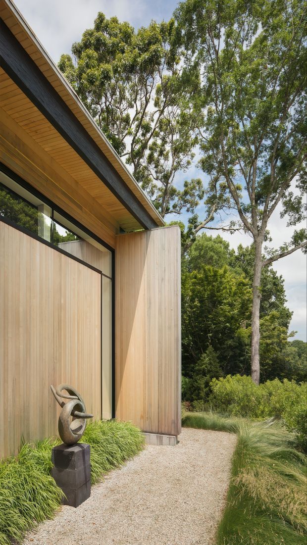 Exterior view of a contemporary low-profile building set within mature landscaped vegetation. The facade is clad in vertically oriented light timber, with large dark-framed glazing and a deep timber-lined roof eave. A pale gravel path curves past ornamental grasses and a small abstract metal sculpture on a dark pedestal.