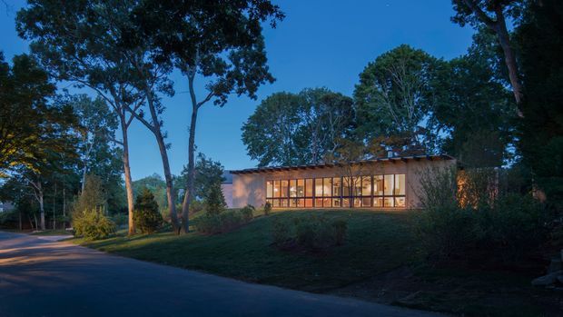 A low, elongated modern house is viewed from a landscaped driveway at dusk. The building has a warm earthy rendered facade, a shallow pitched or low-profile roof, and an expansive glazed frontage glowing with interior light. Mature trees, hedges, and planted garden beds surround the property, creating a secluded suburban or bush-like setting.