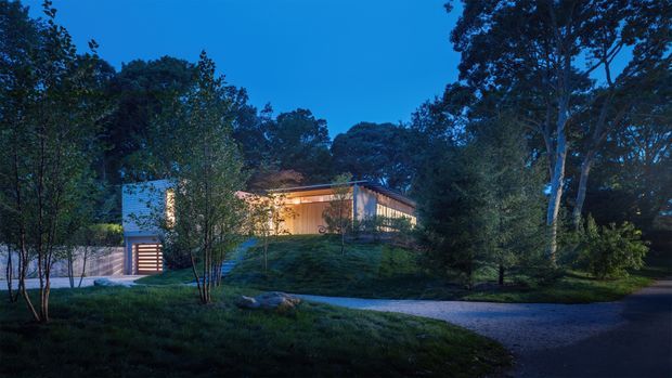 Twilight exterior view of a low-slung contemporary house set within a densely wooded landscape. The home features warm interior lighting, extensive glazing, a flat or gently sloping roofline, pale exterior walls, and an integrated garage or entry structure. A curving gravel driveway and landscaped lawn lead through the trees toward the residence.